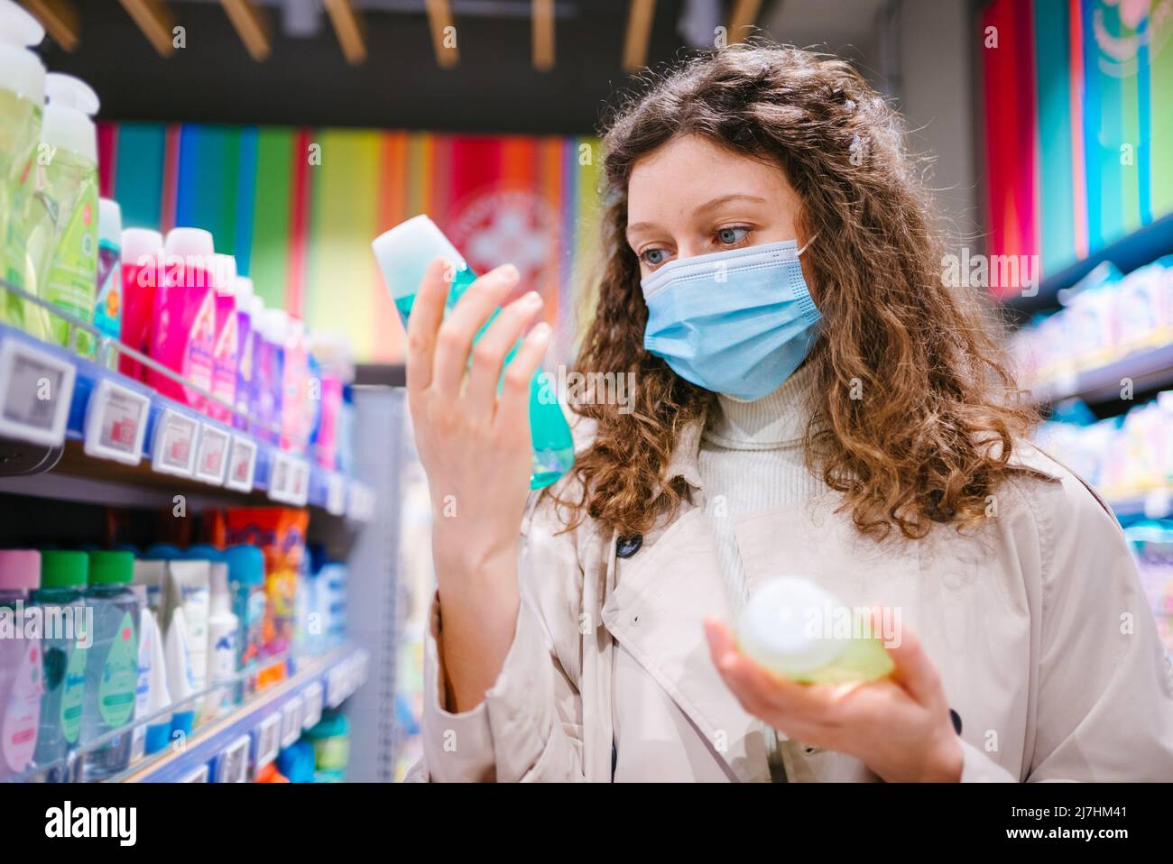Retrato de una mujer compradora con pelo rizado con máscara médica en el supermercado elige champú durante el brote de coronavirus Foto de stock