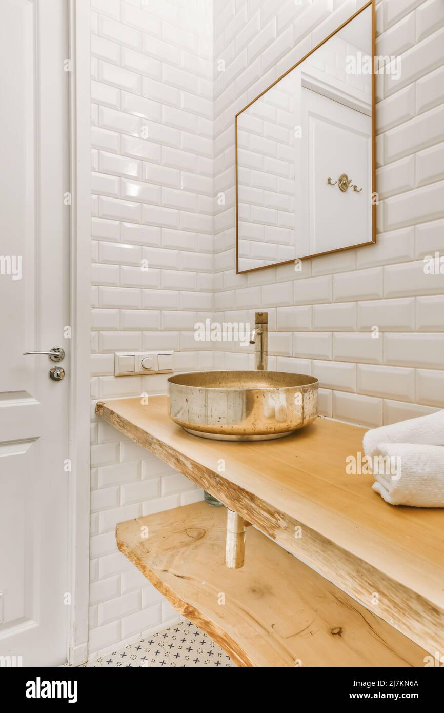 Baño moderno y luminoso con pared de azulejos blancos y espejo colgando sobre un fregadero de cobre sobre una mesa de madera Foto de stock