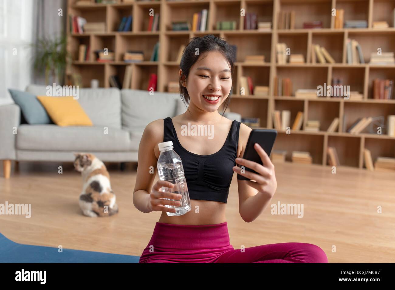 Feliz mujer coreana en ropa deportiva sentada en la colchoneta de yoga en  casa, sosteniendo una botella de agua y usando el teléfono móvil, espacio  de copia Fotografía de stock - Alamy