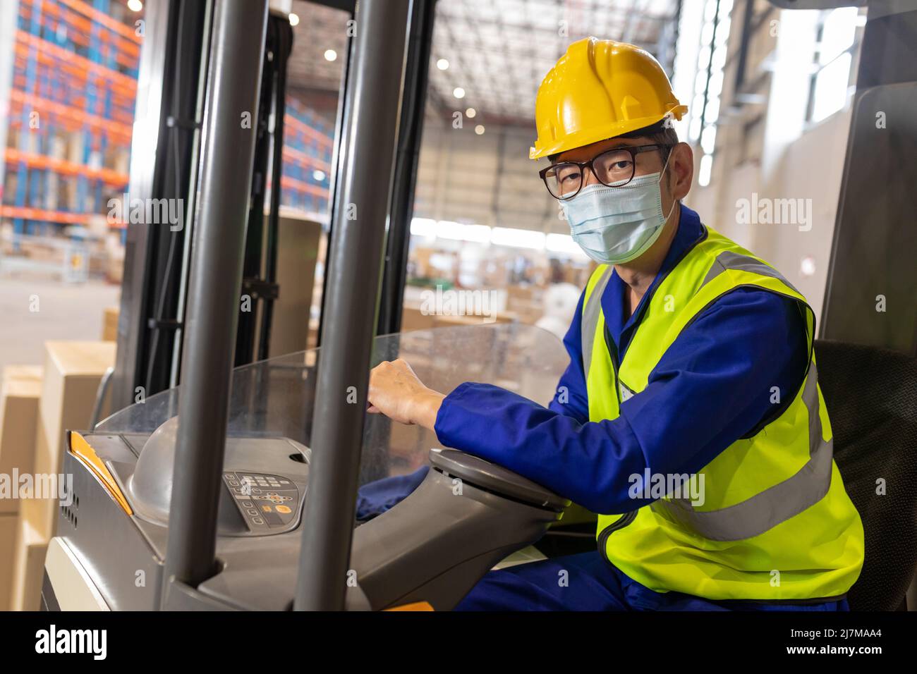 Retrato de trabajador masculino maduro asiático con máscara, chaleco reflectante y casco mientras conduce una carretilla elevadora Foto de stock