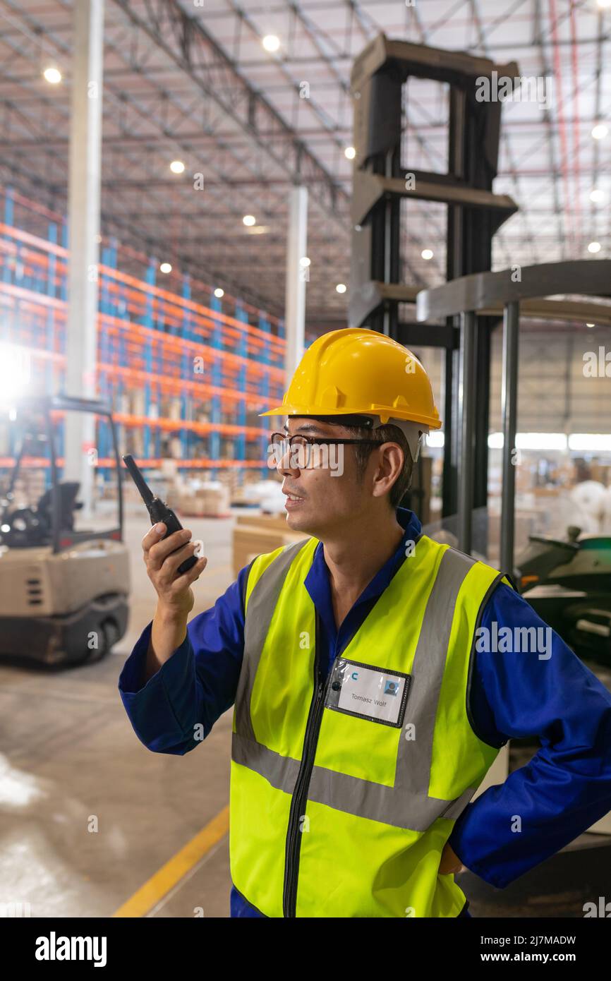 Trabajador de almacén masculino maduro asiático que lleva chaleco reflectante y casco hablando sobre walkie-talkie Foto de stock