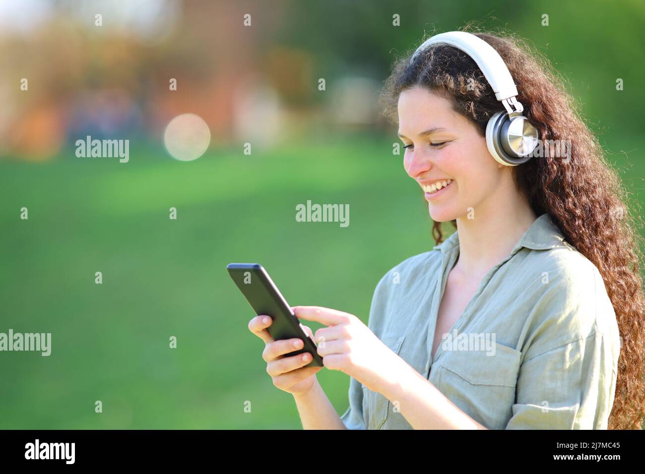 Una mujer feliz escuchando música con auriculares comprueba el smartphone en un parque Foto de stock