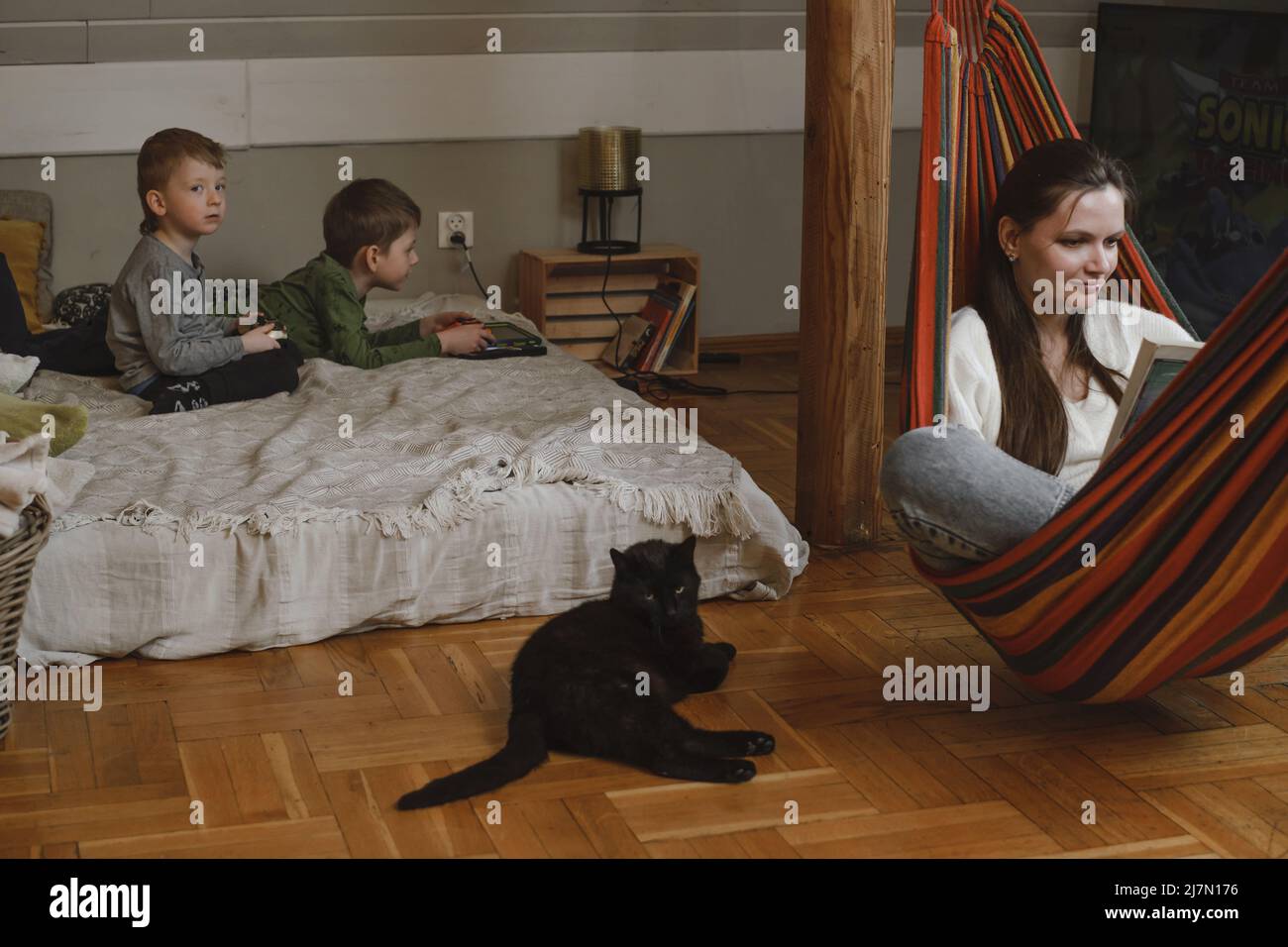 Los niños están jugando a la estación de juegos mientras la madre está leyendo el libro en la hamaca. Familia con gato negro como mascota. Hermanos jugando videojuegos en la cama. Familia Foto de stock