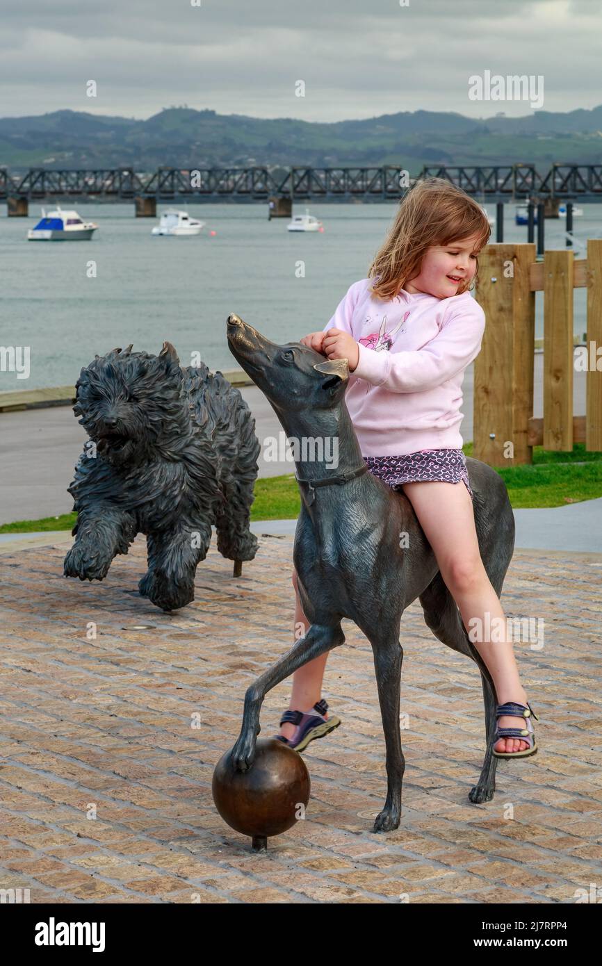 Una chica montando en una de las esculturas de perro 'Hairy Maclary' en la orilla del mar en Tauranga, Nueva Zelanda Foto de stock