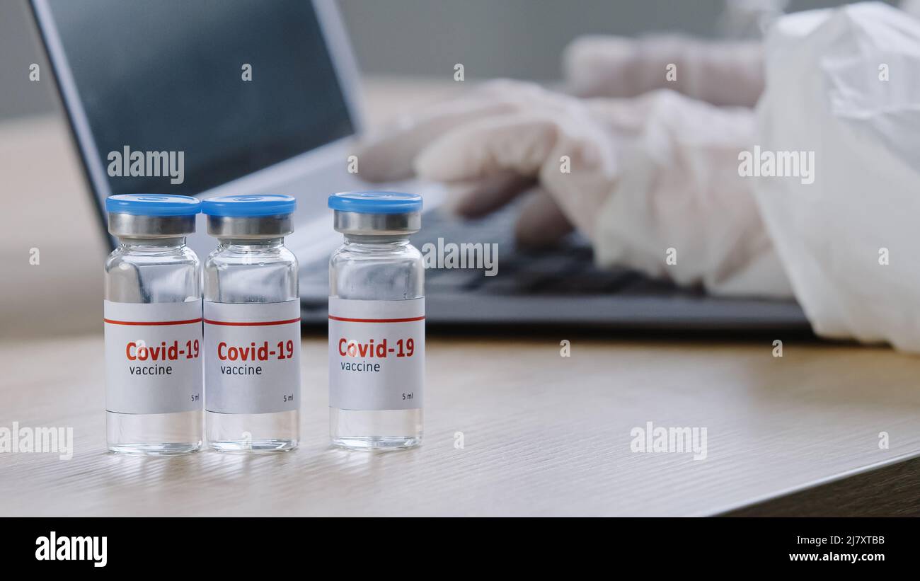 Primer plano de la vacuna covid-19 se encuentra sobre una mesa de madera en la clínica médico inreconocible trabajador científico en guantes de látex mecanografiando en las marcas de los portátiles Foto de stock