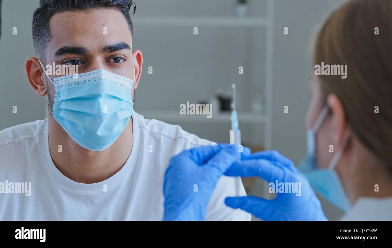 Vista desde detrás de la enfermera del doctor que sostiene la jeringa con la vacuna de la droga para el paciente masculino covid hombre árabe hispano en las manos de las ondas de la máscara no rehúsa Foto de stock