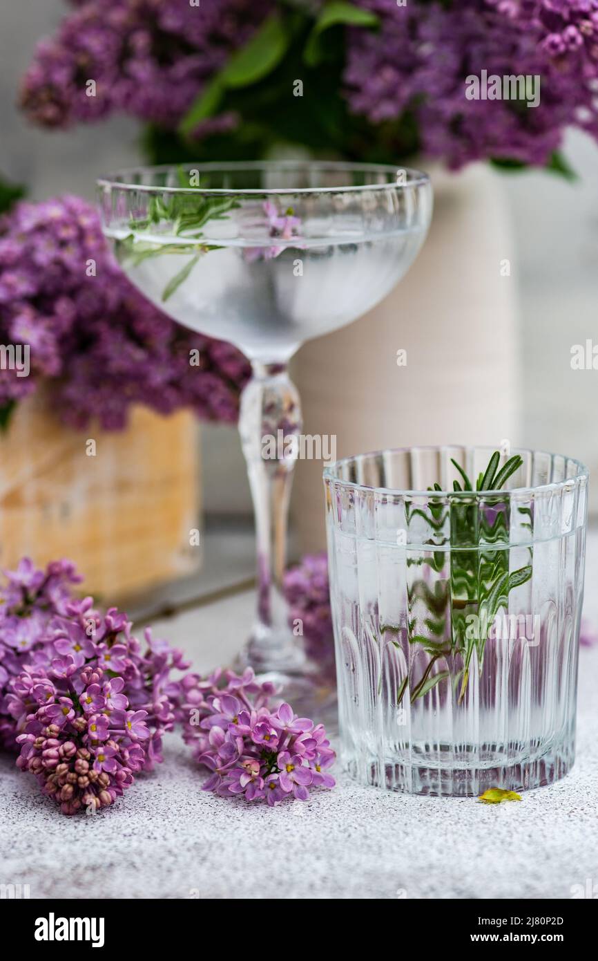 Copa de agua helada y cóctel en un coupé champagne con una ramita de romero y flores lilas Foto de stock