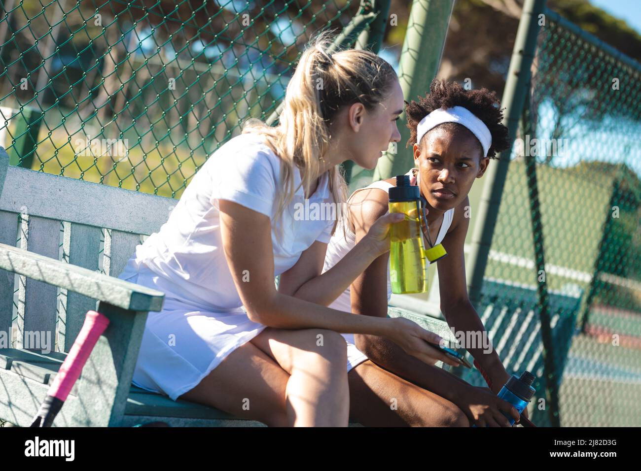 Jóvenes jugadoras de tenis biraciales hablando mientras se sentaban en el banco en la cancha durante el descanso del juego Foto de stock