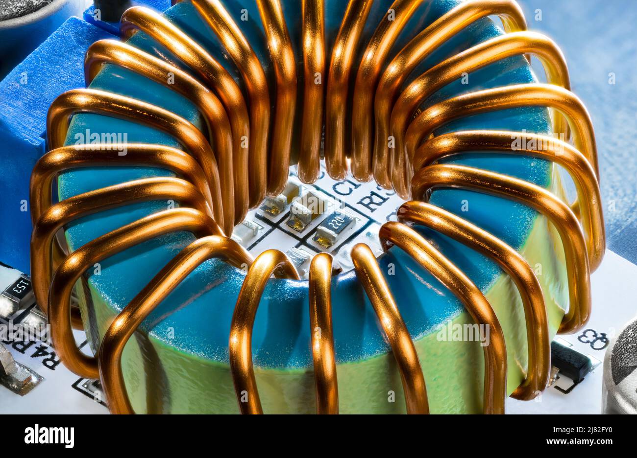 Cierre de bobina de núcleo de ferrita con cable de cobre y resistencias o condensadores en detalle de PCB. Bobina de metal del inductor electrónico toroidal sobre fondo azul. Foto de stock