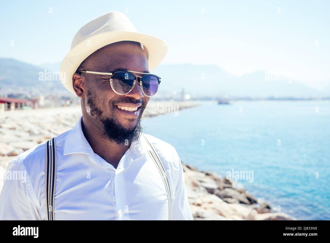 Apuesto african american Person casual moda look conjunto en sombrero de paja, camisa blanca caminando por la costa Tyrkey Foto de stock