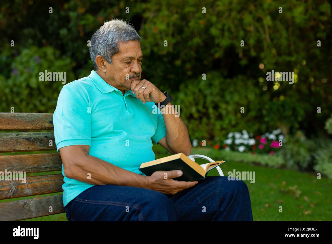 Hombre de edad biracial con mano en libro de lectura de mentón mientras está sentado en banco contra las plantas en el parque Foto de stock