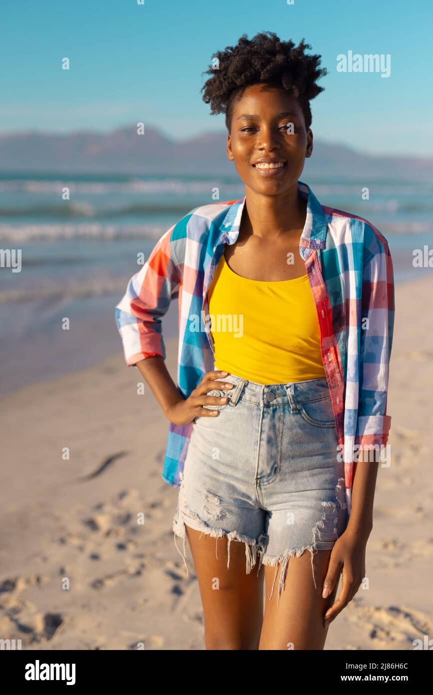 Retrato de una mujer joven afroamericana sonriente con pelo corto de pie en la playa contra el cielo azul Foto de stock