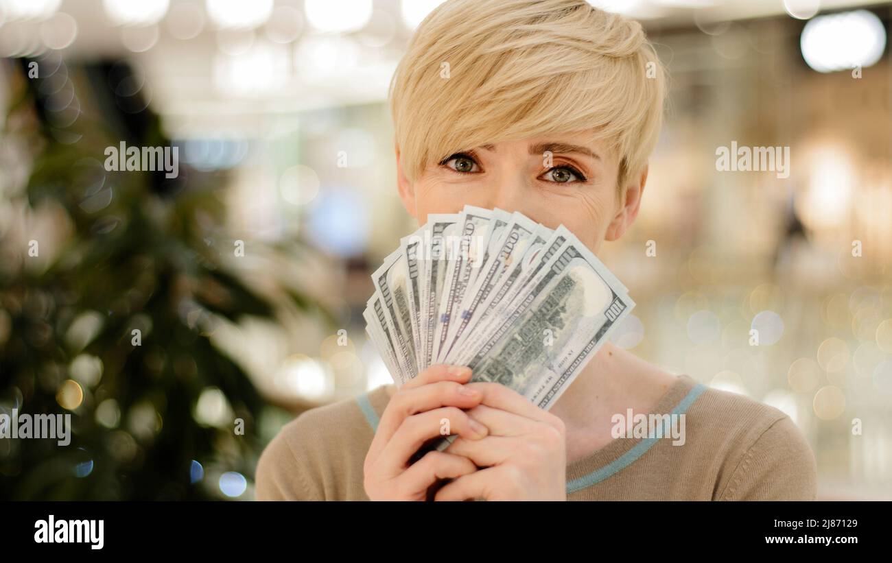 Feliz rica exitosa mujer de negocios caucásica de mediana edad adulta mirando la cámara sosteniendo un ventilador de dinero escondiéndose detrás de billetes de dólar que se muestran Foto de stock