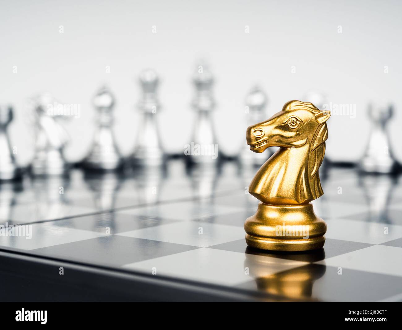 El caballo de oro, la pieza de ajedrez de caballero está sola delante de  piezas de ajedrez de plata sobre tablero de ajedrez sobre fondo blanco.  Liderazgo, luchador, sobreviviente, com Fotografía de