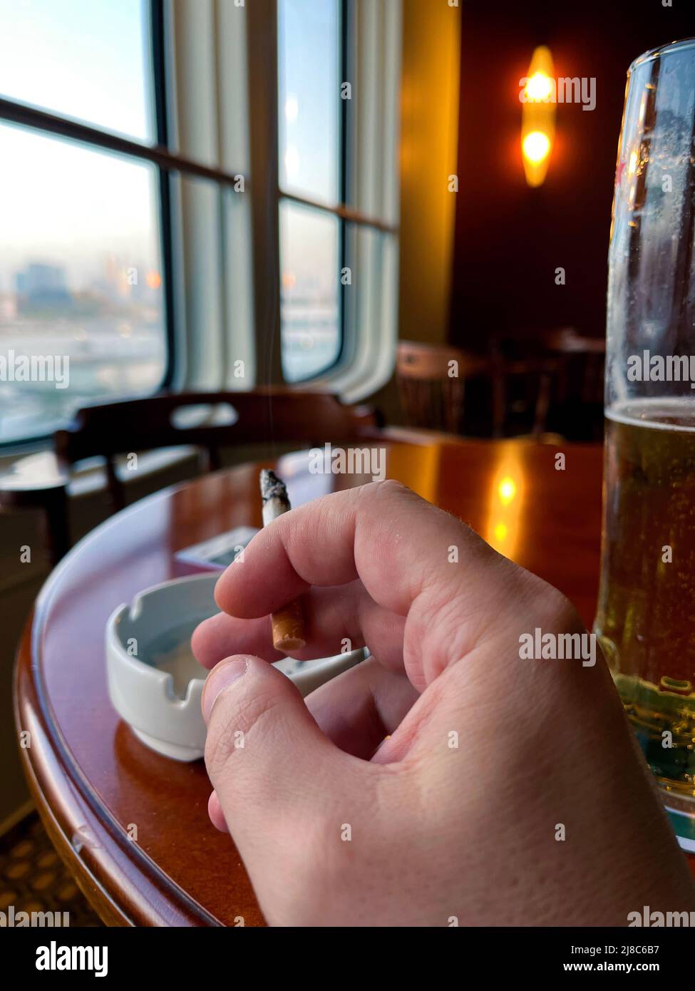 la mano de un hombre sostiene un cigarrillo contra el fondo de las ventanas en una barra de cerveza. el concepto de los daños de fumar. Foto de stock