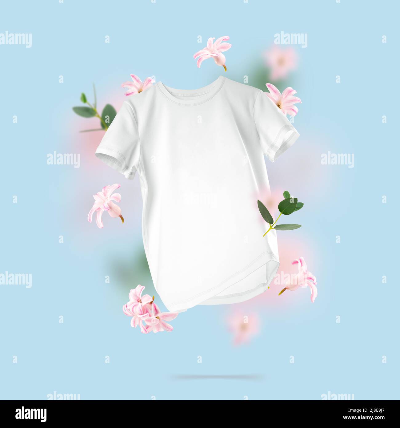 Camiseta blanca de algodón flotando sobre fondo azul. Composición conceptual de una camiseta unisex en blanco y flores en el aire. Vista frontal de la ropa de marca Foto de stock