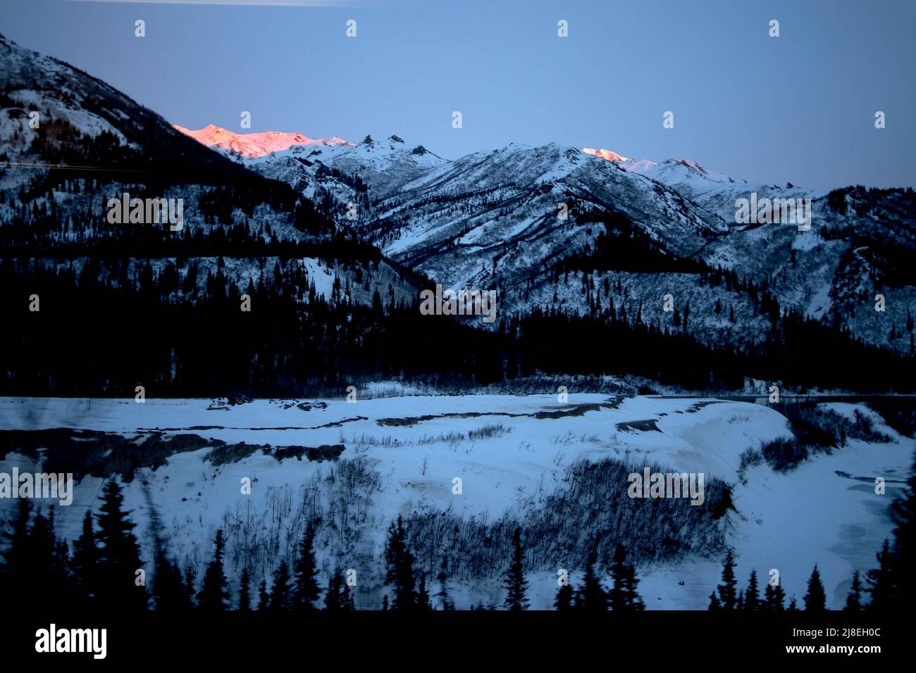 Vista de las montañas y el paisaje nevado, incluyendo un último poco de luz del sol en un acantilado, visto desde la ventana mientras viajaba en un tren de Alaska Railroad. Foto de stock