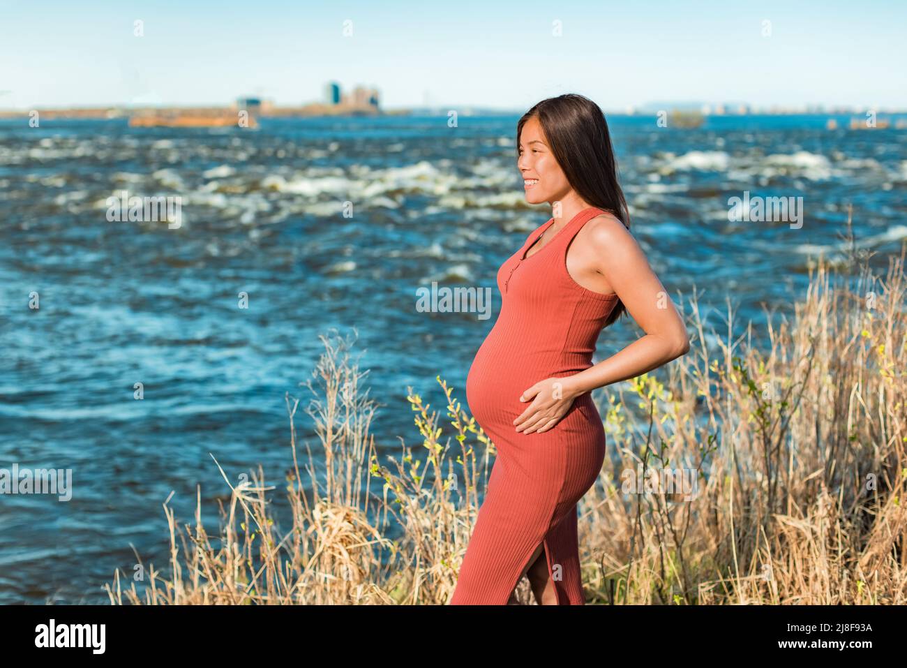 Retrato de maternidad de una mujer asiática durante el embarazo sosteniendo el vientre embarazada contra la naturaleza del fondo del río. Paseo al aire libre en un entorno natural Foto de stock