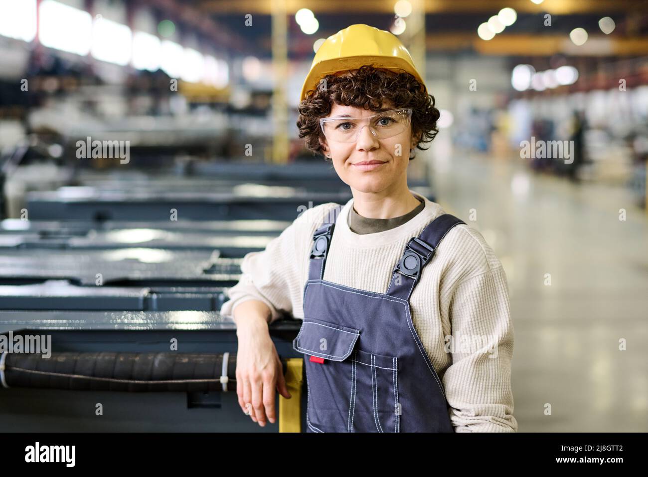 Joven trabajadora feliz de la fábrica moderna que lleva jersey gris, monos  azules y casco amarillo parado por el nuevo equipo industrial Fotografía de  stock - Alamy