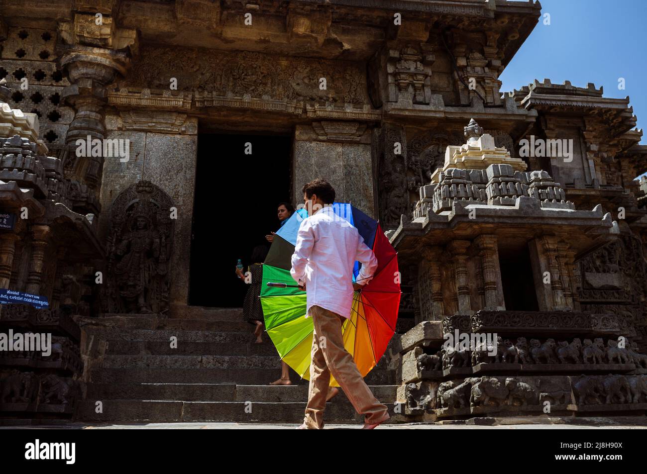 Halebid, Karnataka, India : Un hombre que sostiene un colorido arco iris sol-sombra y su familia visitan el templo Hoysaleswara del siglo 12th Foto de stock