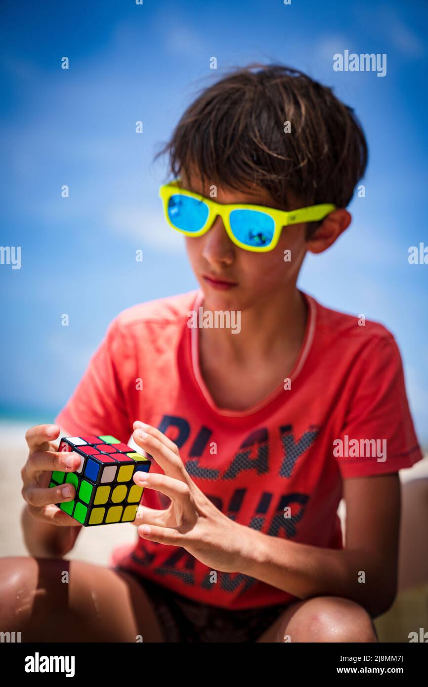 Retrato de un niño con gafas de sol jugando con el cubo de Rubik en una playa, Antigua, Islas de Sotavento, Caribe, Indias Occidentales Foto de stock