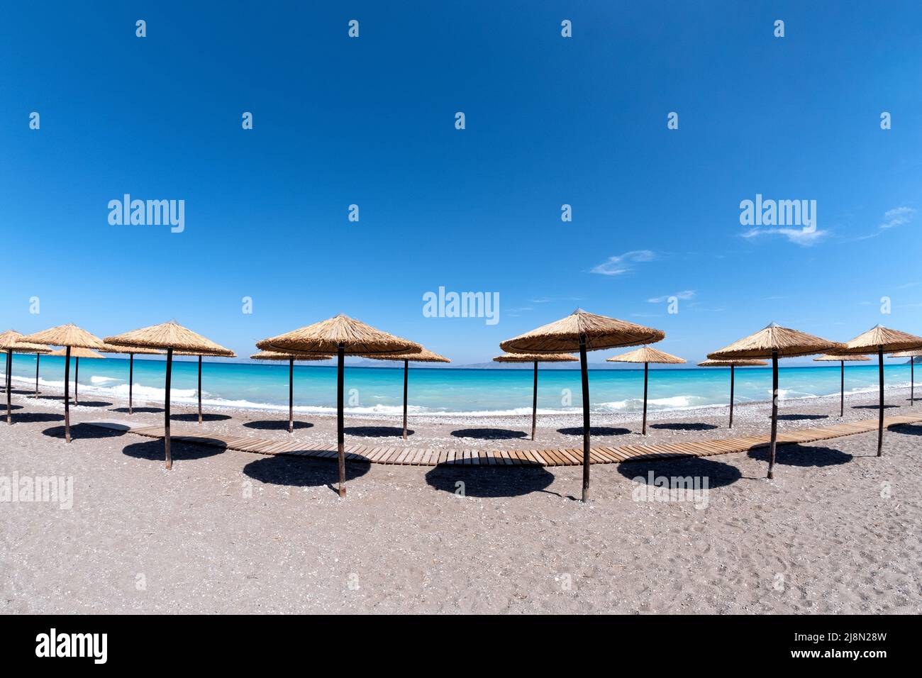 Una hilera de sombrillas de palma o sombrillas de playa de paja bordeadas de gradas filas en una playa de arena vacía con un mar turquesa como telón de fondo y un cielo azul Foto de stock