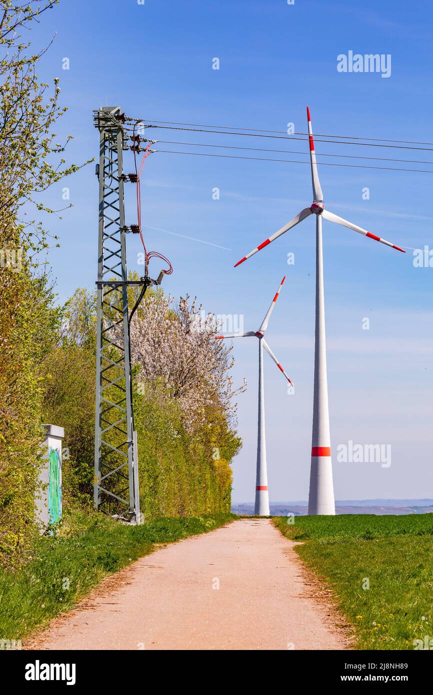 Enormes turbinas eólicas y pilones de electricidad para generar electricidad verde se encuentran junto a un camino de tierra en la Alemania rural Foto de stock