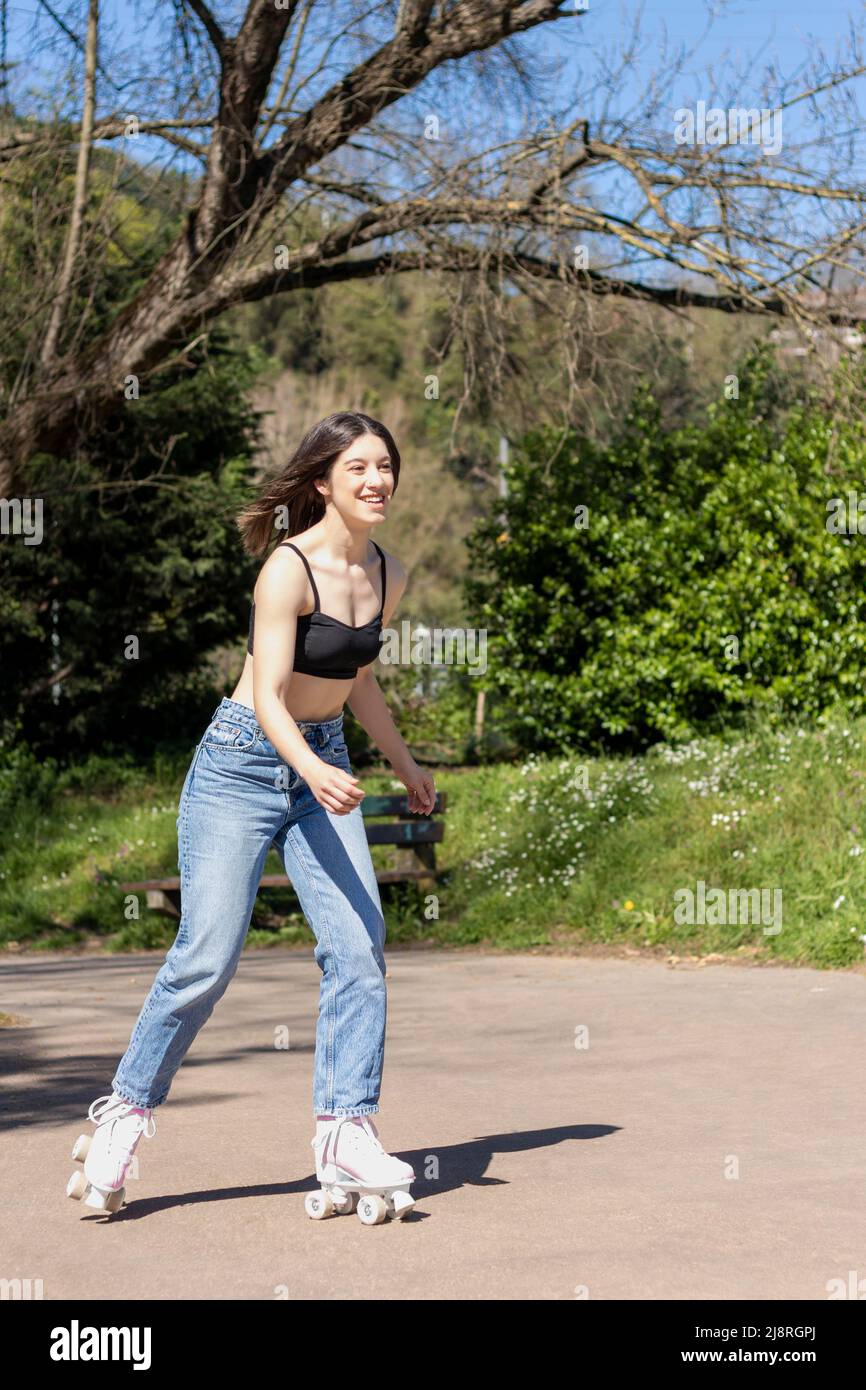 mujer morena patinando en el parque con sujetador deportivo, jeans y patines de roller rosa Foto de stock