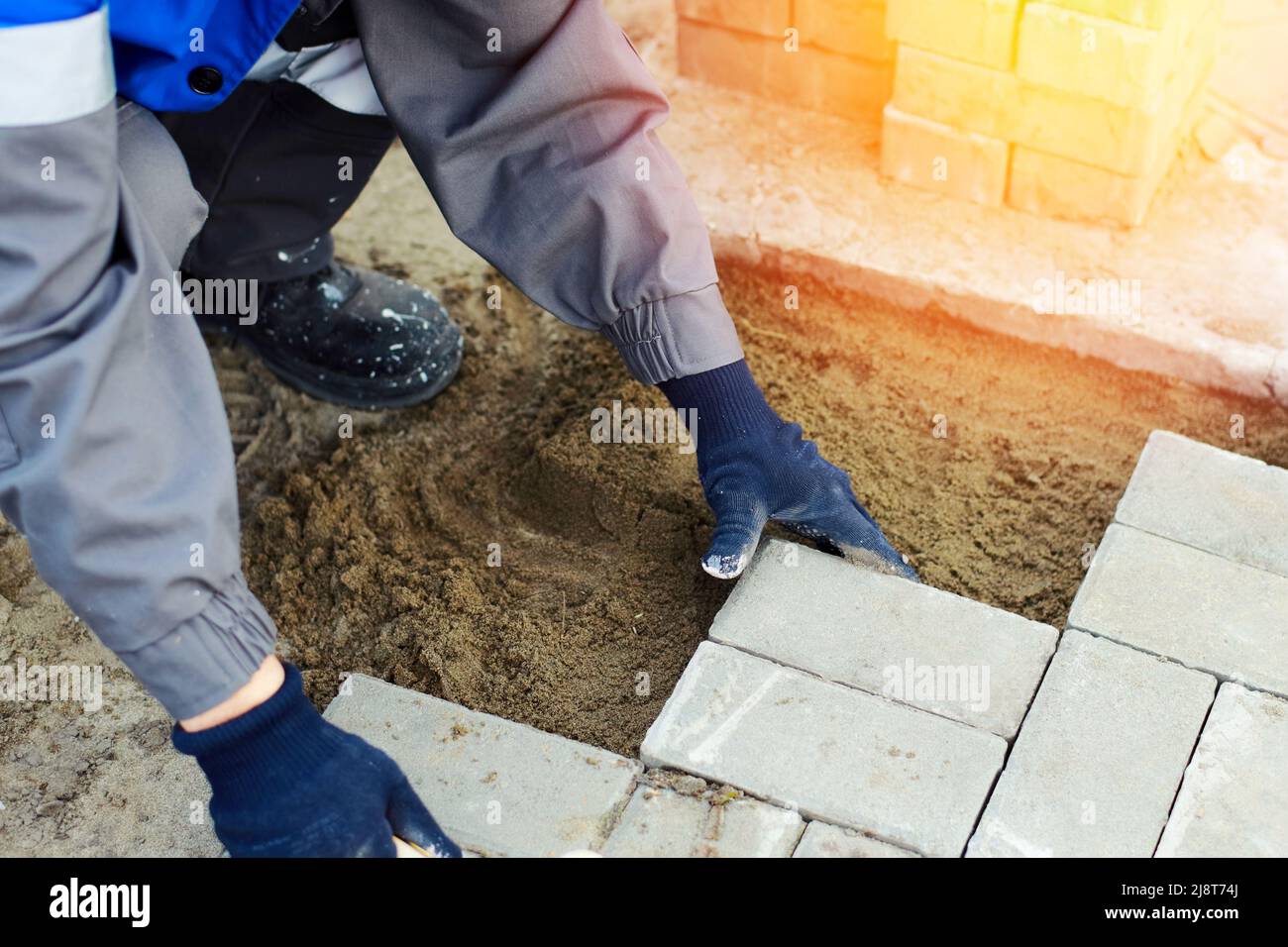 Bricklayer en la ropa de trabajo se sienta en la acera y presenta losas de pavimentación. Vista del trabajador al aire libre. Constructor profesional hace el arreglo del día del verano del territorio. Escena real. Foto de stock