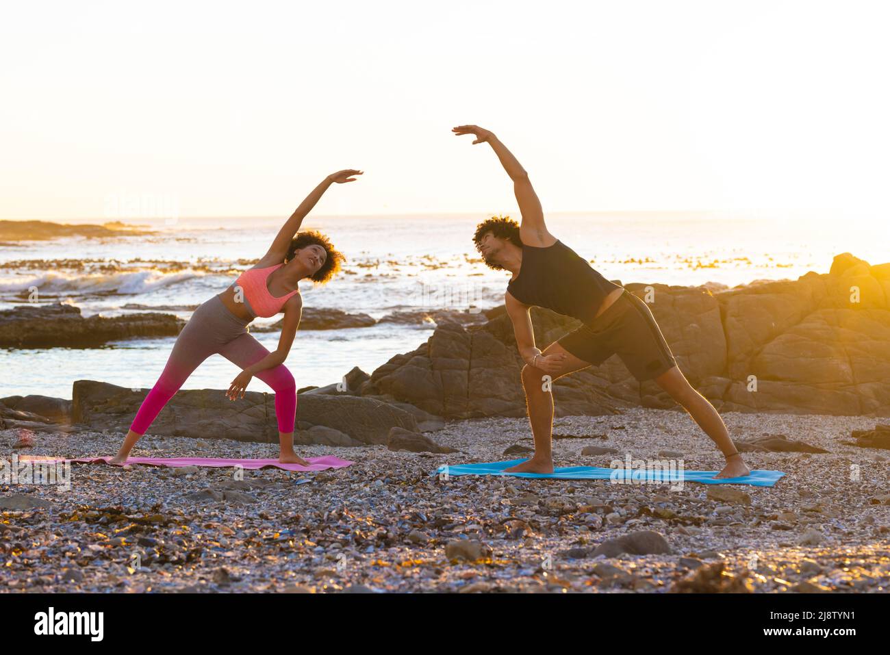 Joven pareja afroamericana doblándose lateralmente mientras hacía yoga en la playa durante la puesta de sol, copiar espacio Foto de stock