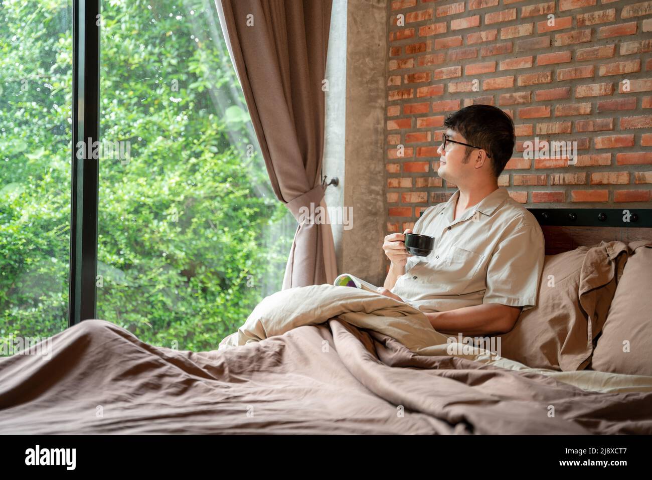 Un hombre sonríe de buen humor por la mañana con una taza de café caliente libro de lectura en el dormitorio de la casa. Foto de stock