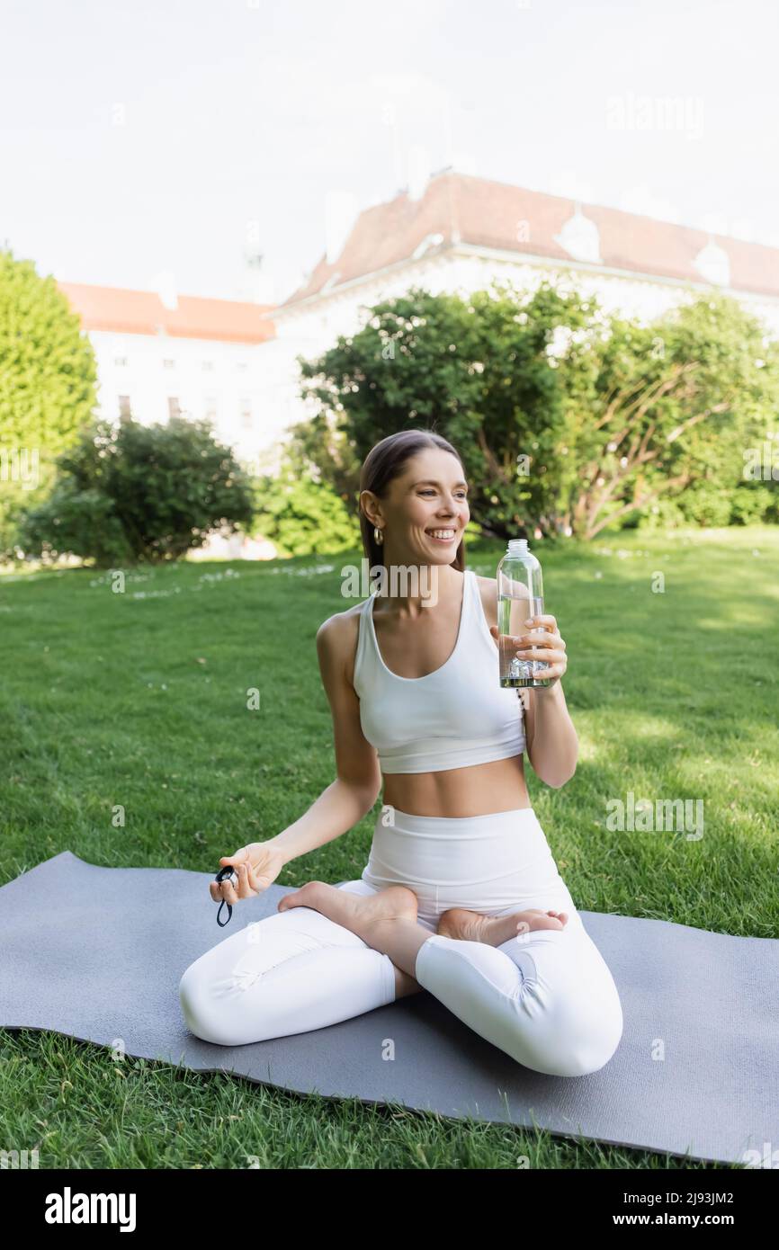 una mujer feliz sentada en lotus pose con una botella de deporte mientras practica yoga en el parque Foto de stock