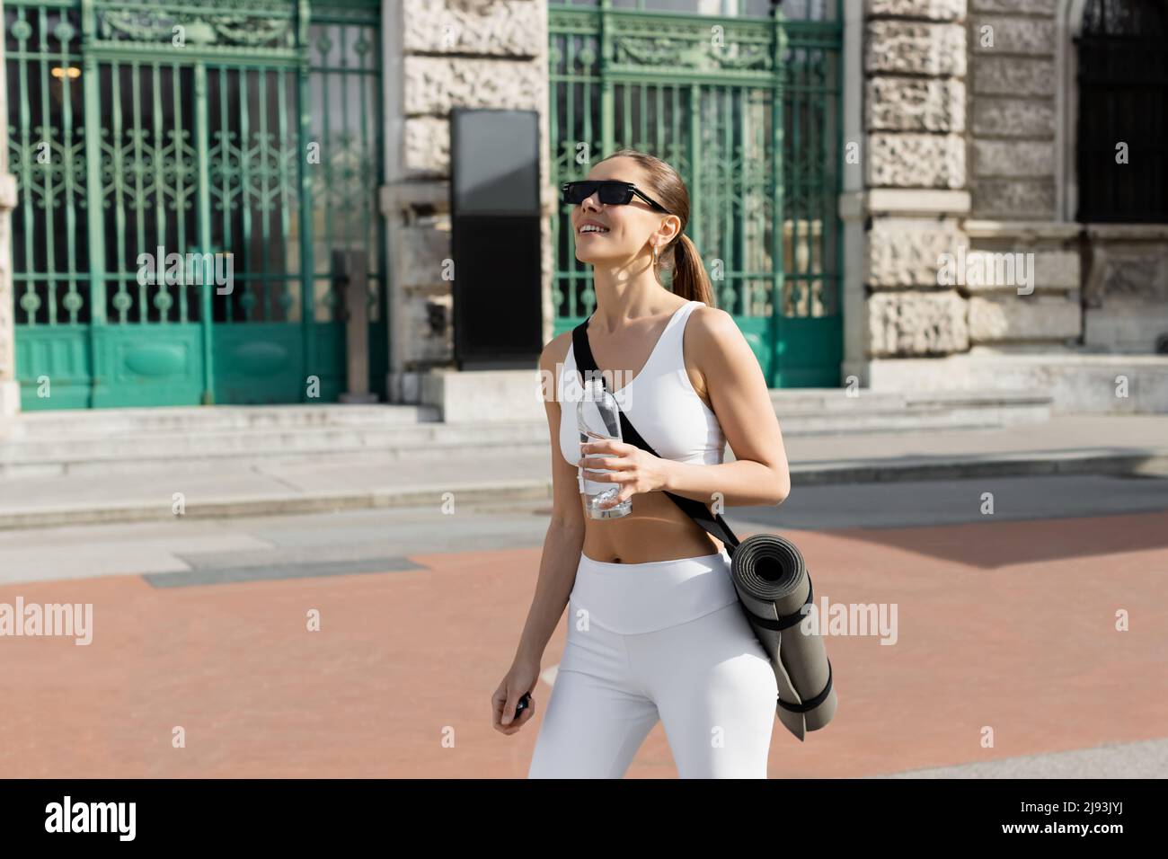 mujer esbelta en ropa deportiva blanca y gafas de sol caminando por la calle con colchoneta de fitness y una botella de deporte Foto de stock