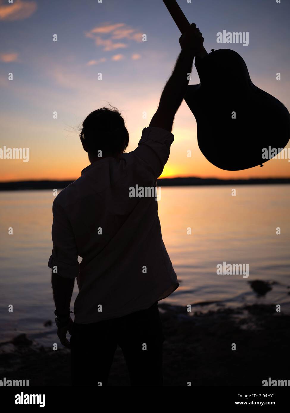 hombre levantando su guitarra en una orilla del lago al anochecer Foto de stock