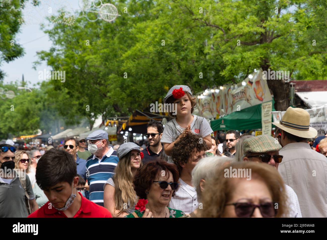 Madrid, España; 15th de mayo de 2022: Un grupo de personas que visitan los  puestos de la feria de San Isidro. En primer plano un niño con una gorra y  claveles en