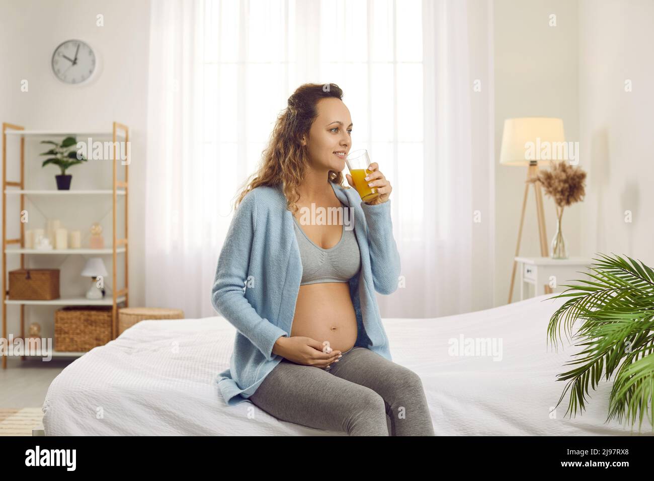 Feliz hermosa mujer embarazada saludable sentada en la cama y bebiendo jugo de naranja Foto de stock