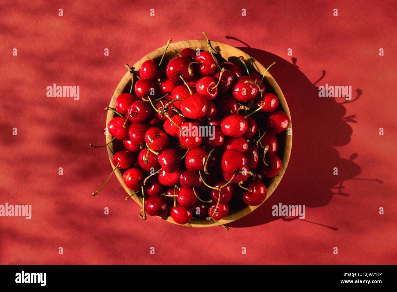 Vista superior de cerezas dulces maduras en un tazón de madera bajo la luz del sol sobre un fondo rojo. Foto de stock
