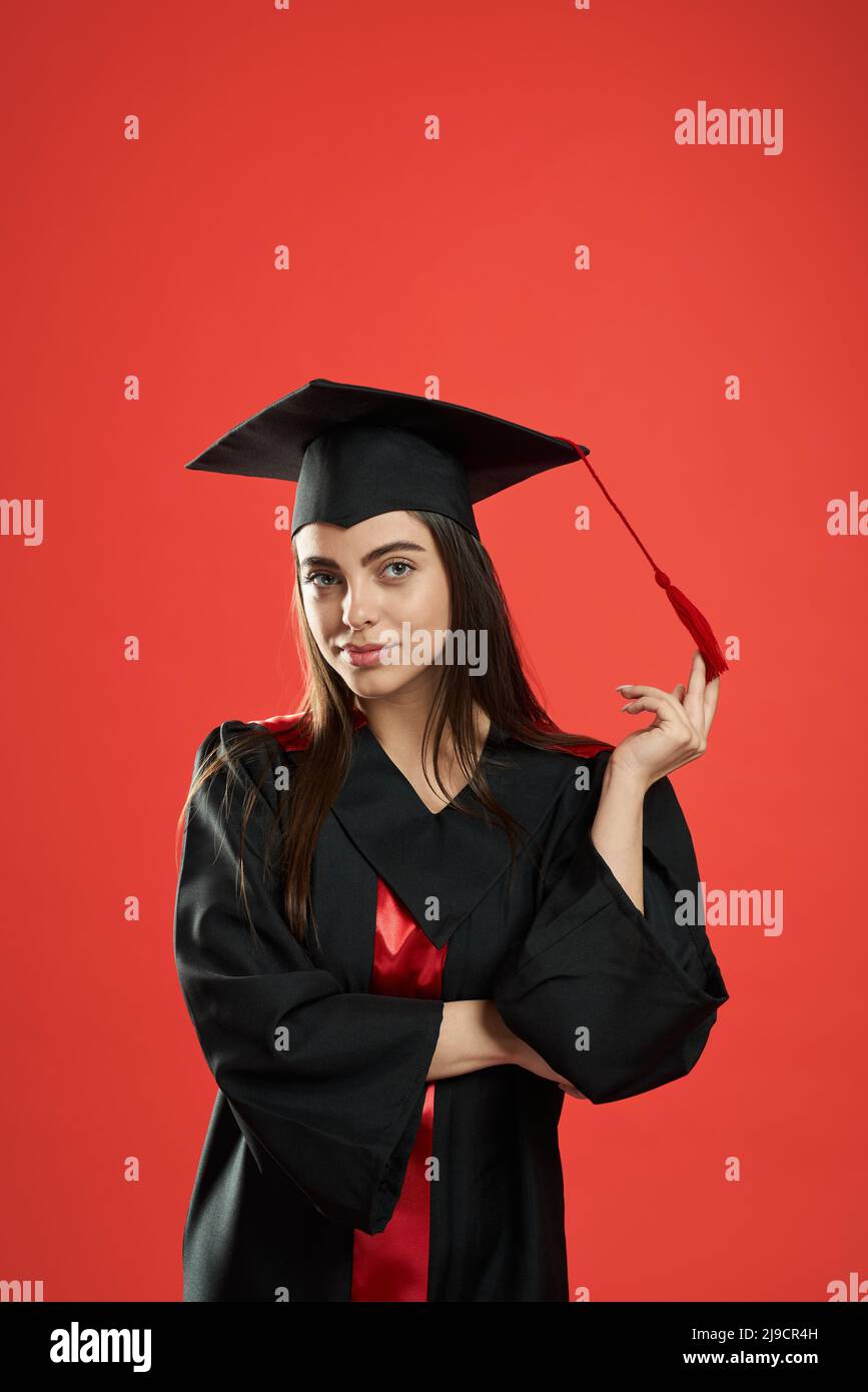 Vista frontal de la chica bonita en mortarboard y vestido de posgrado de pie con las manos cruzadas. Una estudiante muy femenina sonriendo, mirando la cámara. Aislado sobre fondo de estudio rojo, Foto de stock