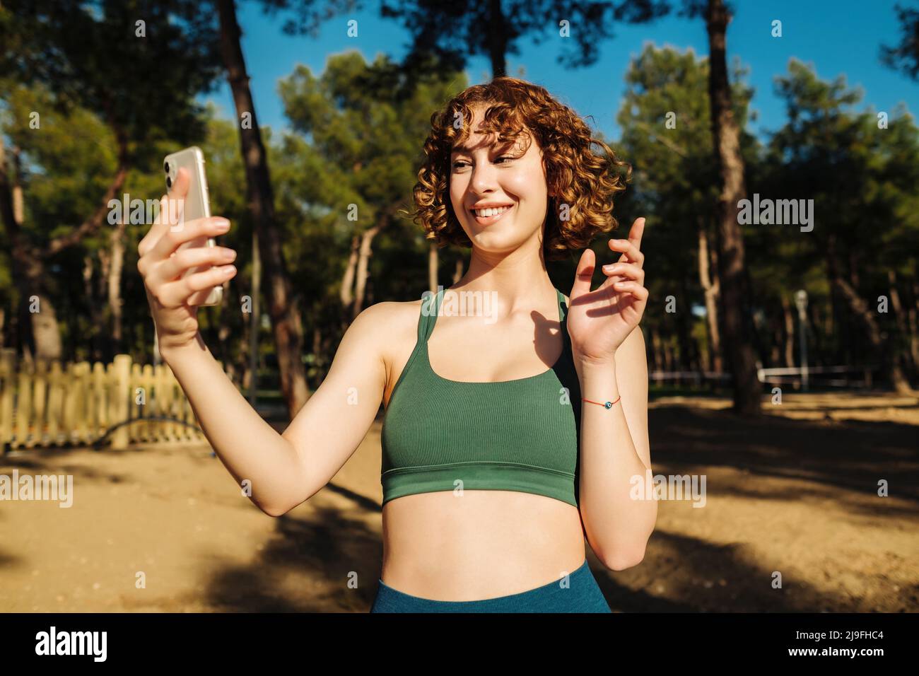 Mujer joven pelirroja usando sujetador deportivo en el parque de la ciudad,  al aire libre sosteniendo el teléfono mirando la pantalla agitando la mano  video que llama a distancia amigo en línea