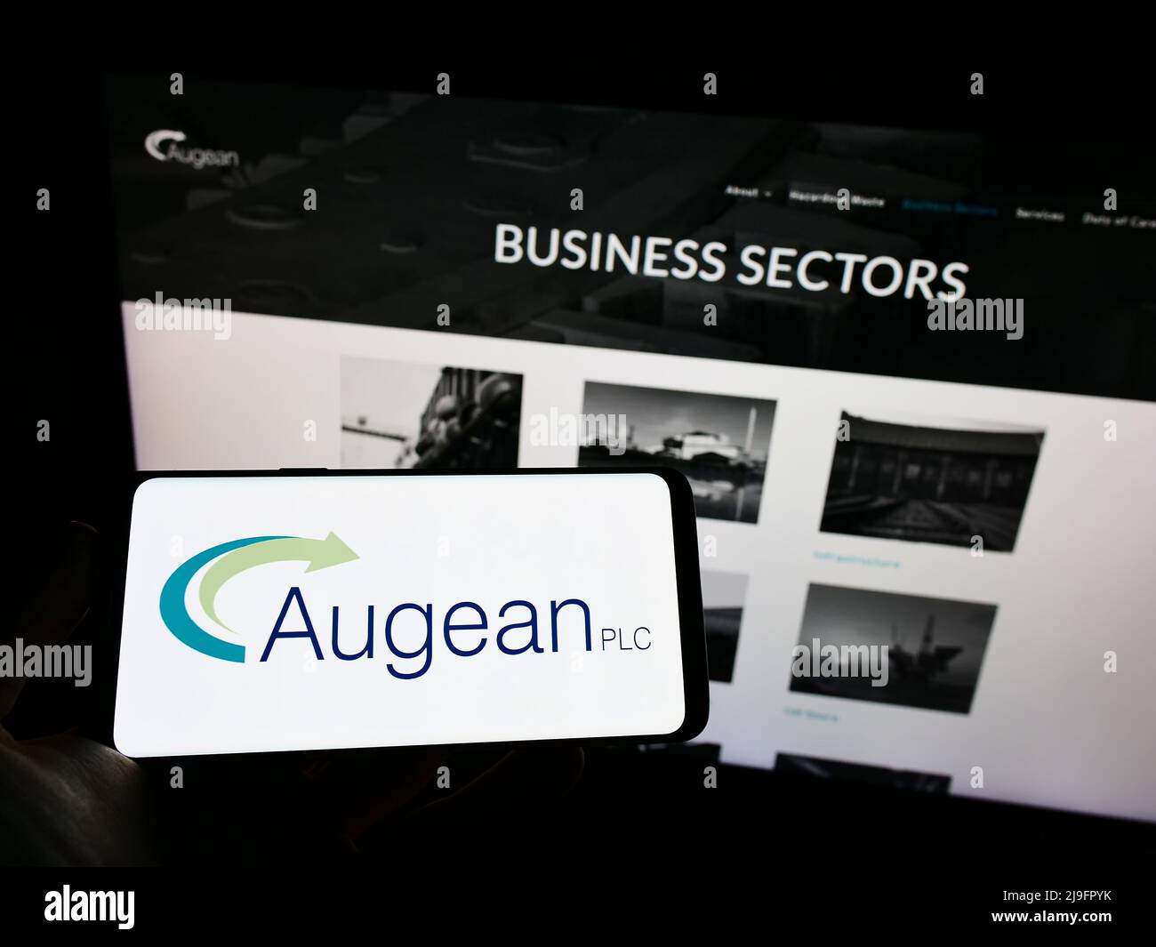 Persona que sostiene un teléfono móvil con el logotipo de la empresa británica de gestión de residuos Augean plc en la pantalla frente a la página web de la empresa. Enfoque en la pantalla del teléfono. Foto de stock