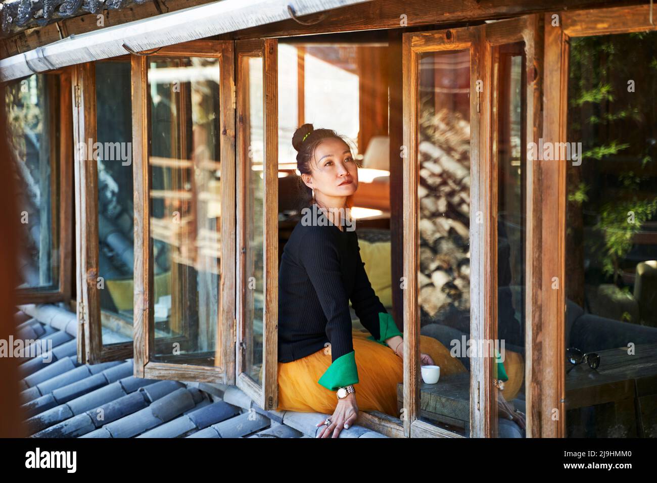 mujer asiática sentada en el alféizar de la ventana disfrutando de una taza de café Foto de stock