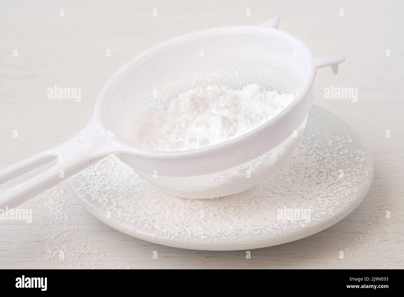 Azúcar glaseado en un tamiz de plástico sobre una placa de cerámica blanca sobre madera blanca. Foto de stock