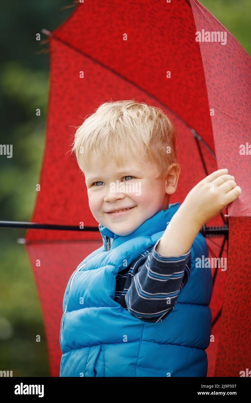 Niño tomando paraguas rojo con cara feliz sonriente. Foto vertical. Foto de stock