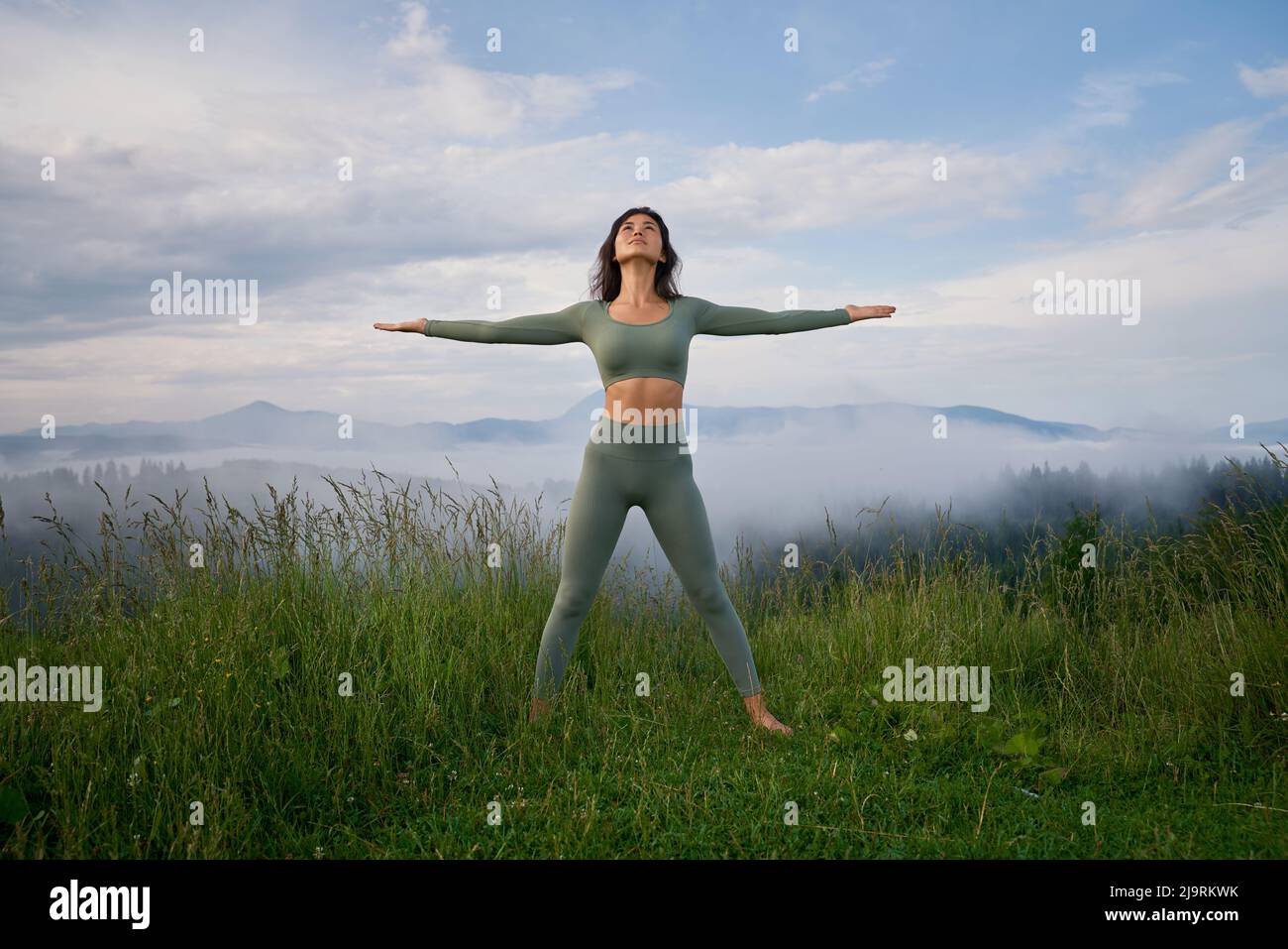 Vista frontal de una mujer morena de pie sobre hierba en la colina de la montaña en pose de ganador, mirando hacia arriba. Atractiva mujer haciendo yoga al aire libre por la mañana entre la niebla. Concepto de salud. Foto de stock