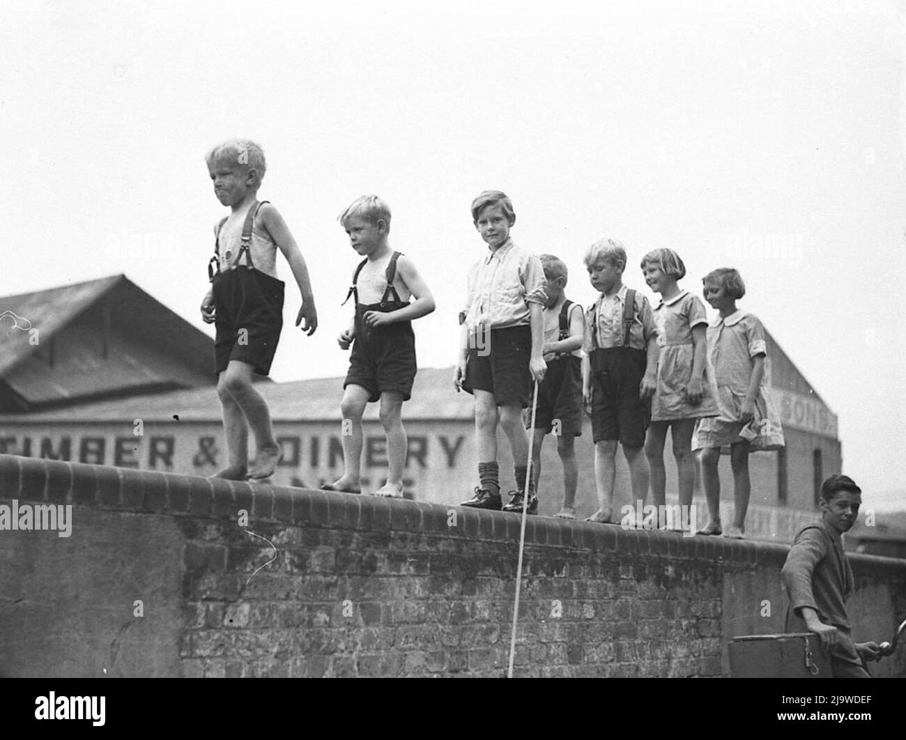 Sam Hood - Cinco niños pequeños y dos niñas caminando a lo largo de una pared de ladrillo - 1935 Foto de stock