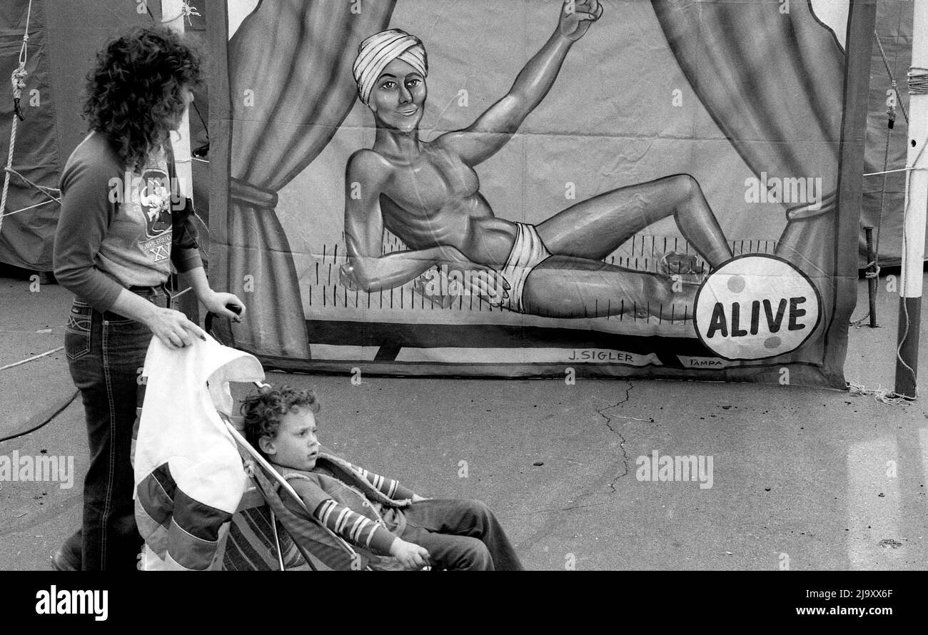 Una mujer con un niño en un cochecito pasa por un cartel de circo que representa a un hombre tumbado en una cama de clavos. Foto de stock