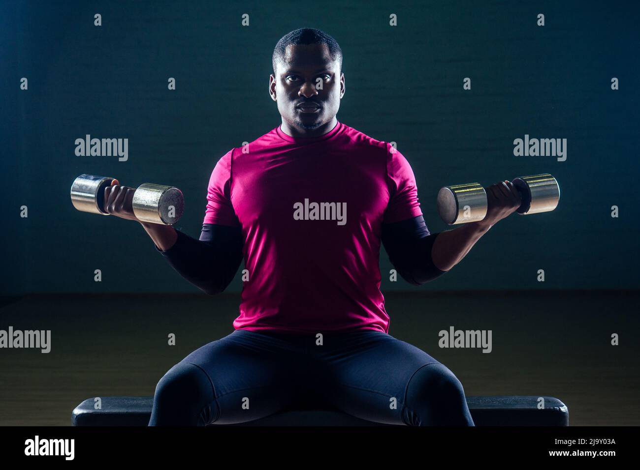 hombre afroamericano musculoso haciendo ejercicios de flexiones con pesas en el gimnasio sobre un fondo negro Foto de stock