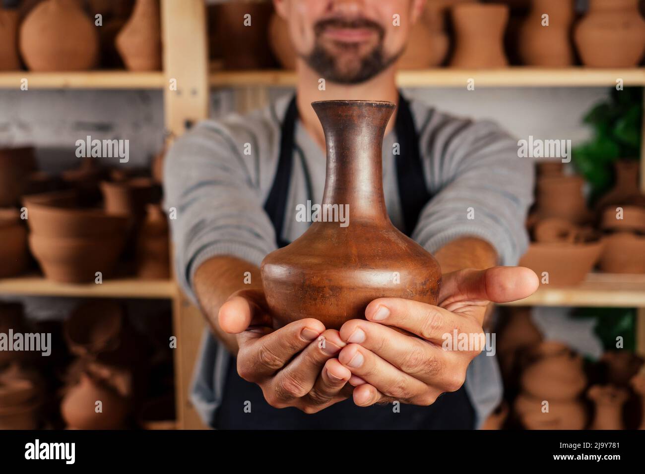 El maestro alfarero en delantal esculpe un tazón profundo de arcilla marrón en una rueda de alfarero en el taller sobre el fondo de estantes con platos Foto de stock