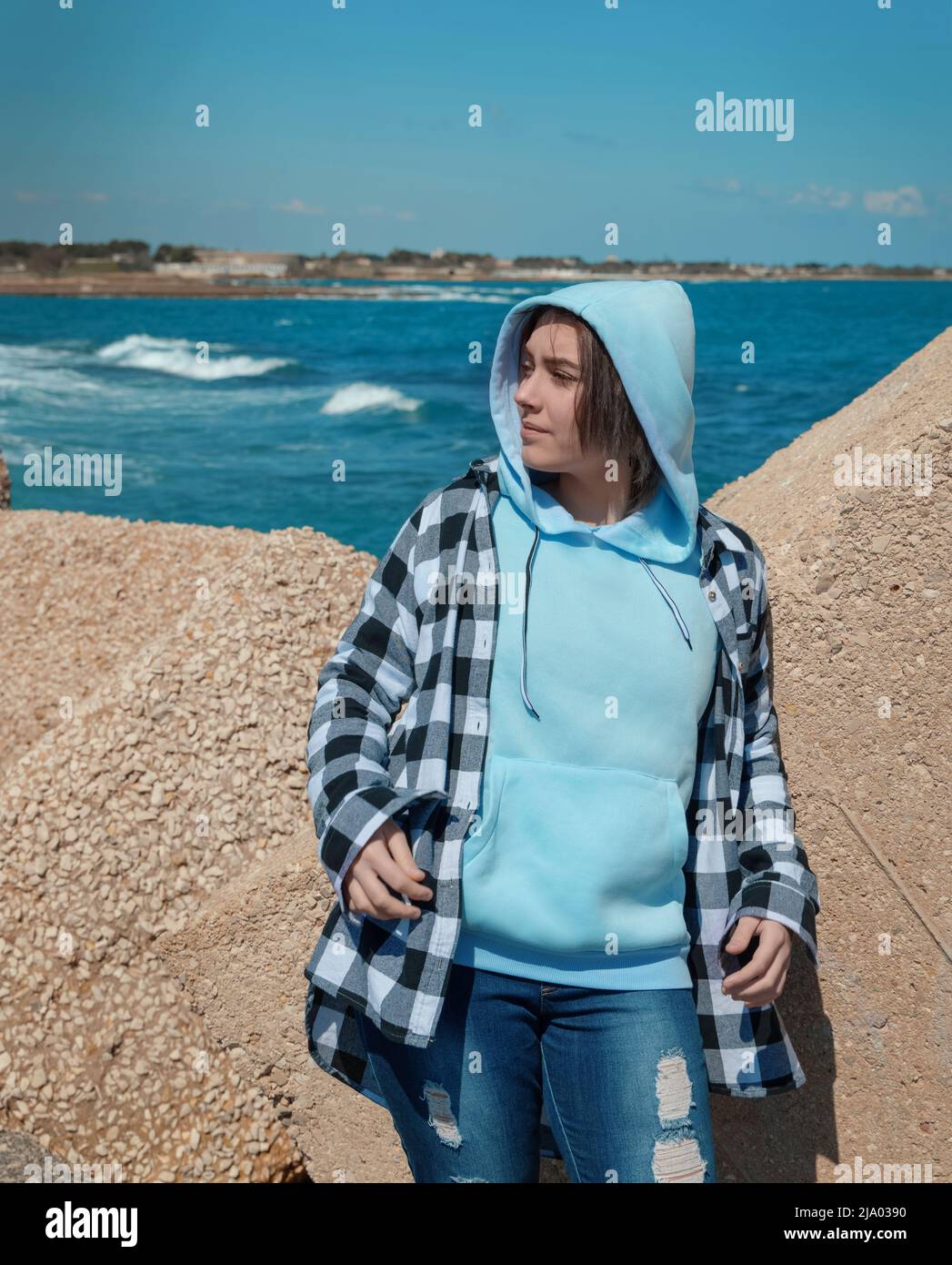 Adolescente en sudadera azul claro con capucha y camisa a cuadros mirando a  un lado. Adolescente permanece al aire libre cerca del mar, bloques de  hormigón y cielo azul, día ventoso. Ropa