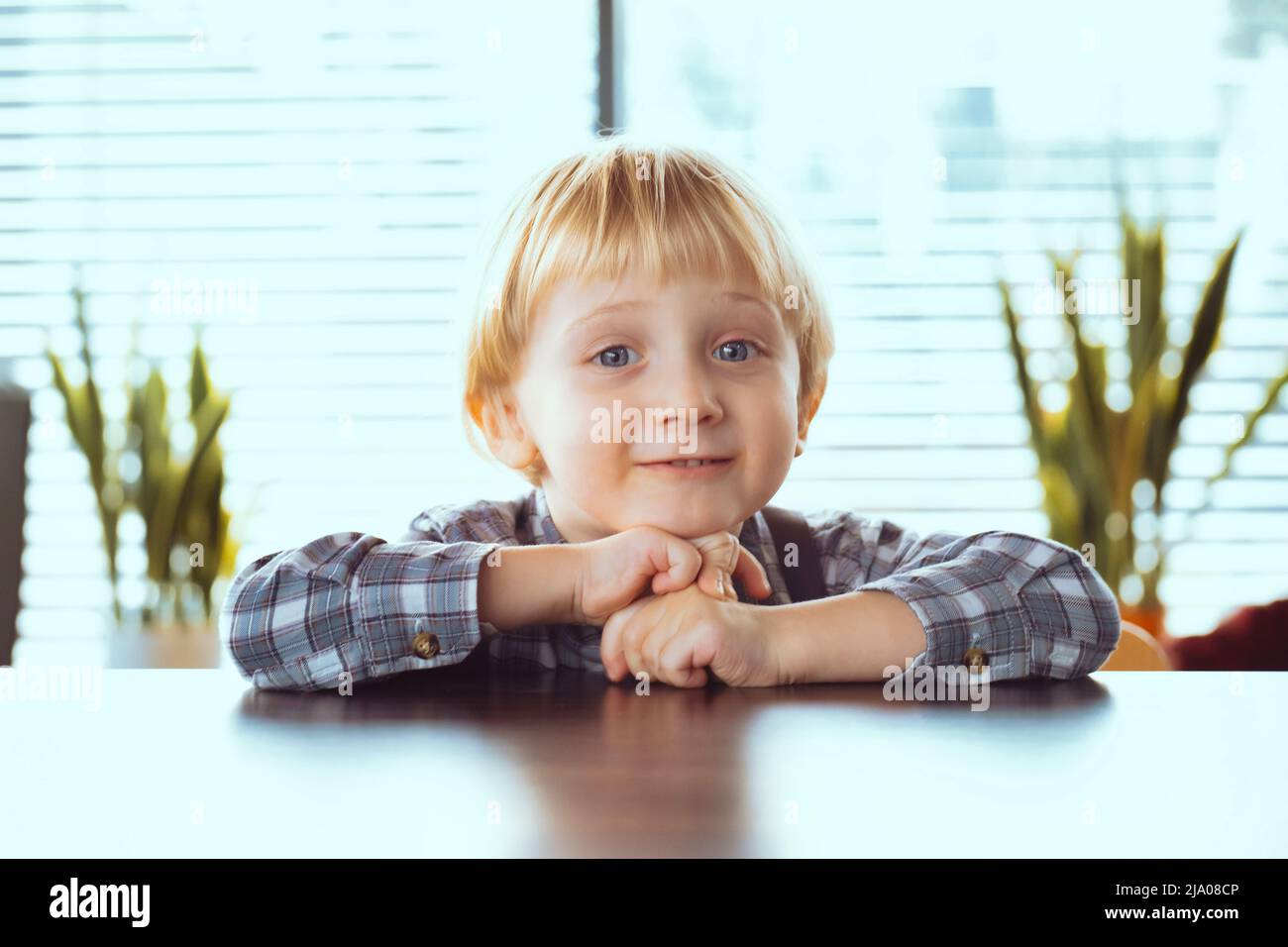 Un niño de 3 años con pelo rubio en camisa sonríe. Un niño feliz en la mesa. Foto de stock